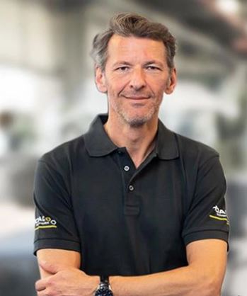 Ein lächelnder Mann in einem schwarzen Polohemd mit gefalteten Armen vor verschwommener Kulisse.
